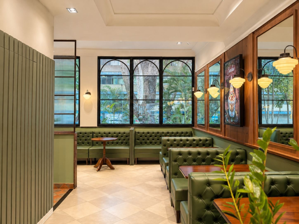 Salão de restaurante com sofás capitonê verdes, painéis de madeira, espelhos, arandelas e janelas em arco voltadas para as árvores