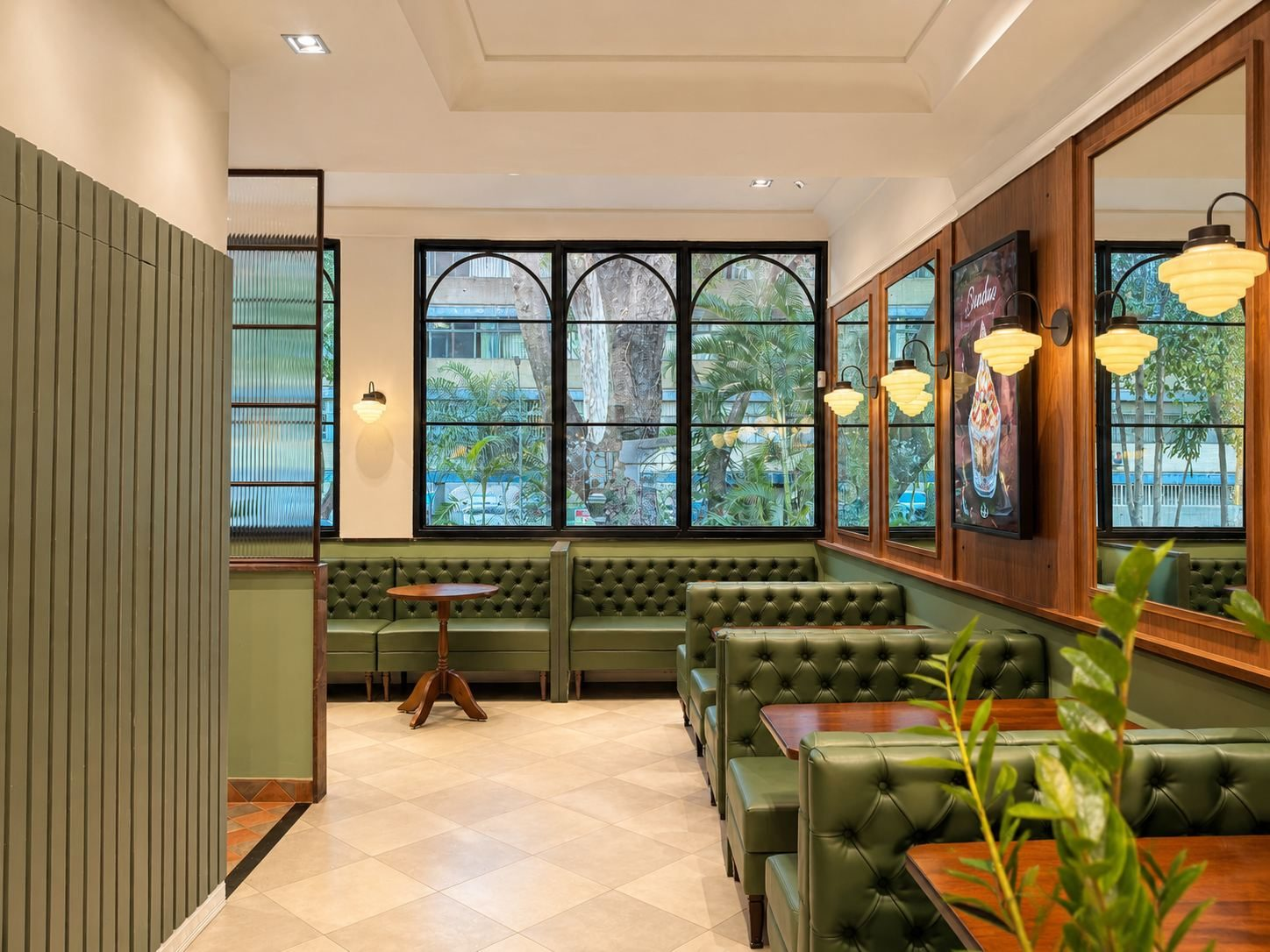 Salão de restaurante com sofás capitonê verdes, painéis de madeira, espelhos, arandelas e janelas em arco voltadas para as árvores