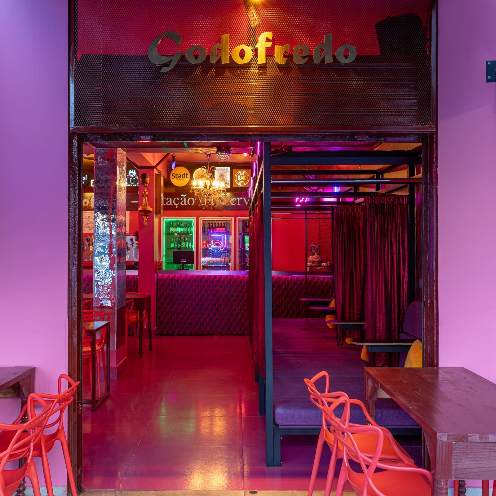 Fachada do bar Godofredo, com luz rosa e vermelha, cadeiras vermelhas na área externa, balcão capitonê e cabines com cortinas